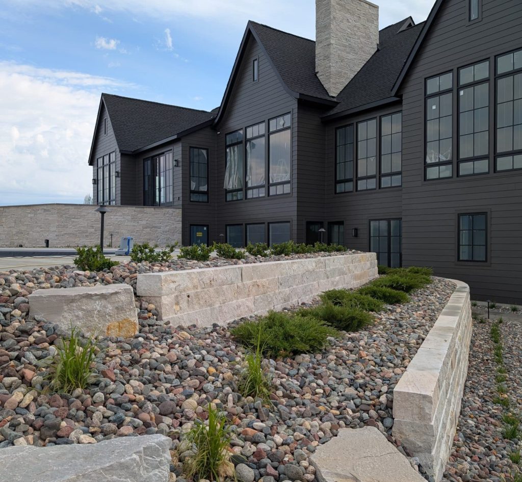 A multi-tiered stone block retaining wall surrounded by river rock mulch and low evergreen shrubs in front of a modern dark charcoal grey house.